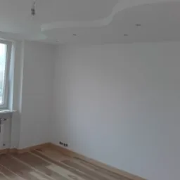 Jasne panele ułożone w pokoju z białymi ścianami i ozdobnym sufitem podwieszanym z oświetleniem punktowym, widoczne gniazdka elektryczne przy podłodze.