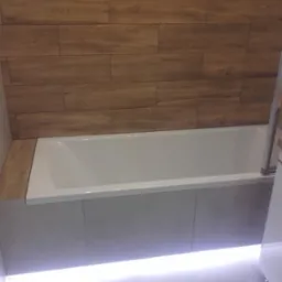 Nowoczesna biała wanna z obudową, podświetlona listwą LED, z drewnopodobnymi płytkami na ścianie w łazience.