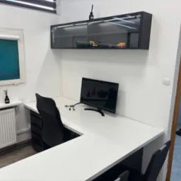 Białe biurko narożne z czarnym krzesłem obrotowym i monitorem, nad nim szafka wisząca z przeszkleniem i oświetleniem LED, wewnątrz modele ciężarówek i koparki.