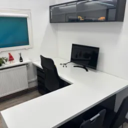 Białe biurko narożne z czarnym krzesłem biurowym na tle okna z roletą i szafki wiszącej z zabawkami, minimalistyczne wnętrze biura.