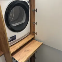 Pralka i suszarka w zabudowie meblowej z drewnianymi elementami i wysuwanym blatem. Białe fronty szafek z widocznymi zawiasami. Nowoczesne rozwiązanie do małej łazienki.