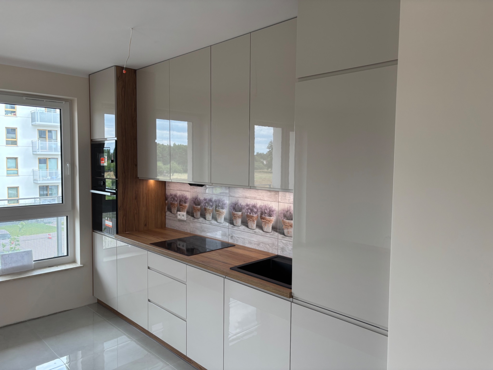 Nowoczesna kuchnia z białymi, lakierowanymi frontami, drewnianym blatem i panelem z motywem lawendy. Widok na okno i zabudowę piekarnika.
