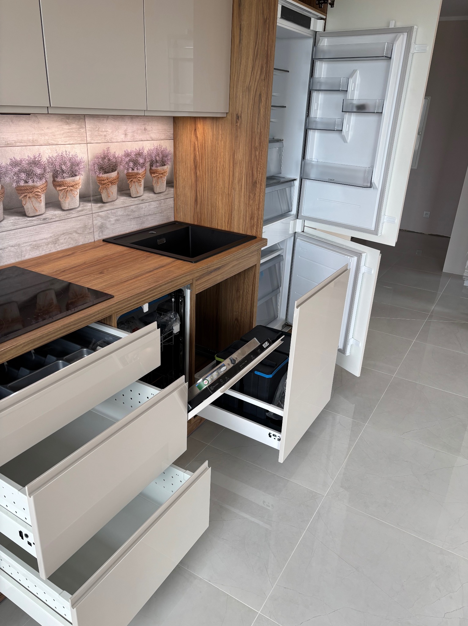 Nowoczesna kuchnia z drewnianym blatem, czarnym zlewem i otwartymi szufladami. Widoczna zintegrowana lodówka i zmywarka. Na ścianie dekoracyjny motyw z lawendą.