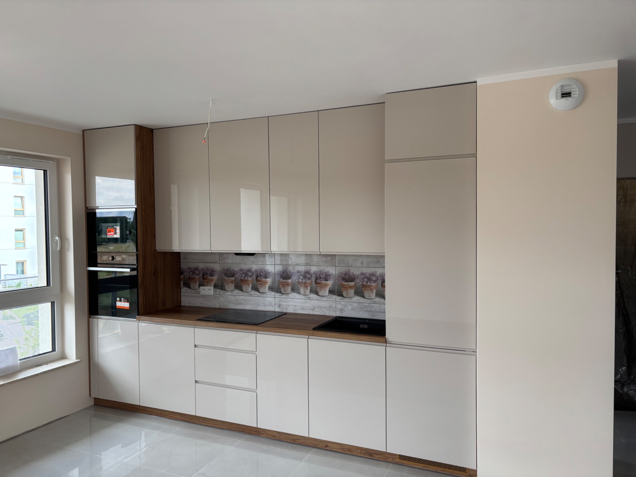 Nowoczesna kuchnia w beżowych odcieniach z połyskującymi frontami, drewnianym blatem i płytkami z motywem lawendy nad blatem roboczym. Widoczne sprzęty AGD.