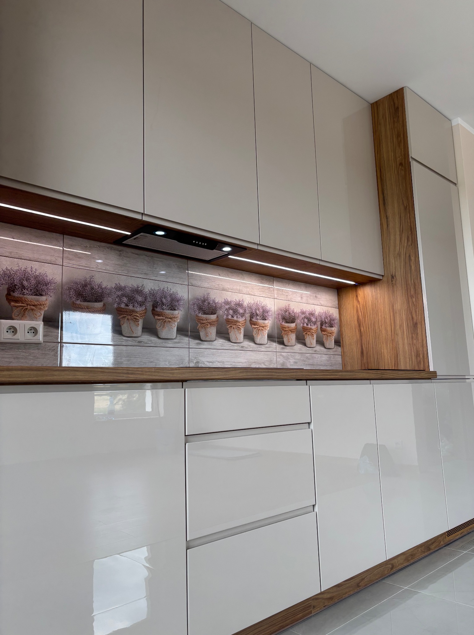 Biała, nowoczesna kuchnia z połyskiem. Nad blatem rząd doniczek z lawendą na płytkach. Oświetlenie LED pod szafkami. Elementy wykończenia w kolorze drewna.
