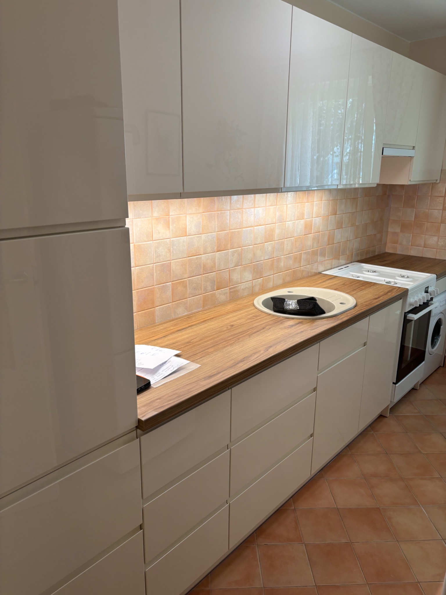 Jasna kuchnia z drewnianym blatem, beżowe płytki na ścianie i brązowa podłoga. Na blacie leżą dokumenty i telefon. W zlewie czarna ścierka.