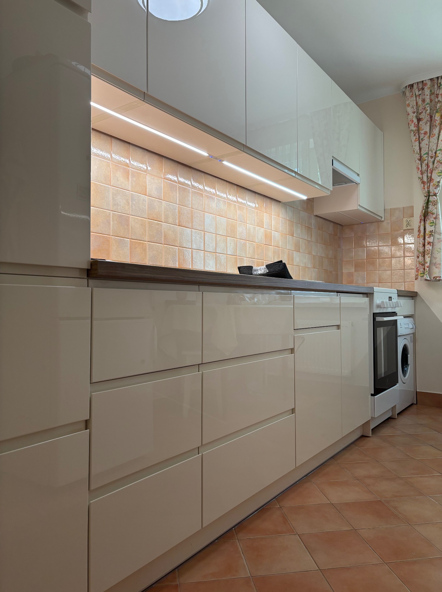 Jasna kuchnia z połyskiem, oświetlenie LED pod szafkami wiszącymi, brązowe płytki na ścianie i podłodze, pralka w zabudowie. Ujęcie z niskiej perspektywy.