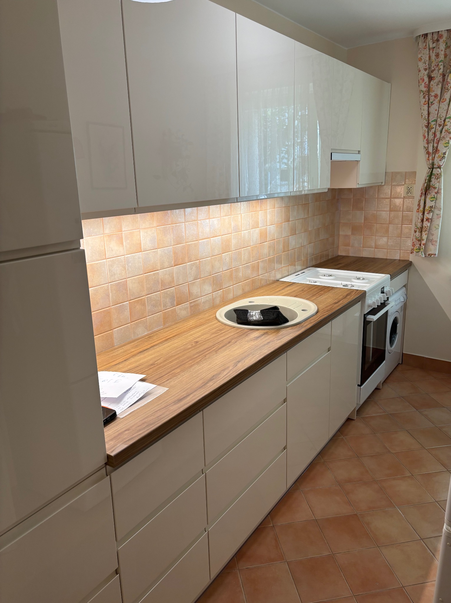 Biała, błyszcząca kuchnia na wymiar z drewnianym blatem w Radomsku. Uwagę zwraca nietypowe połączenie pralki i kuchenki obok zlewu. Jasne płytki na ścianie i podłodze.