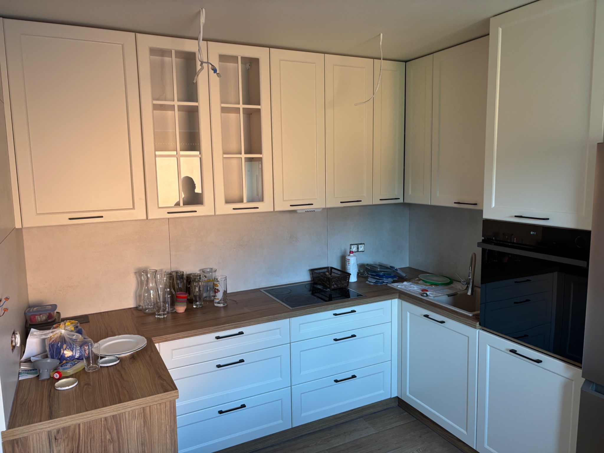 Jasna kuchnia z białymi szafkami i drewnianym blatem. Na blacie akcesoria kuchenne i szklane naczynia. Widoczne niedokończone podłączenia elektryczne.