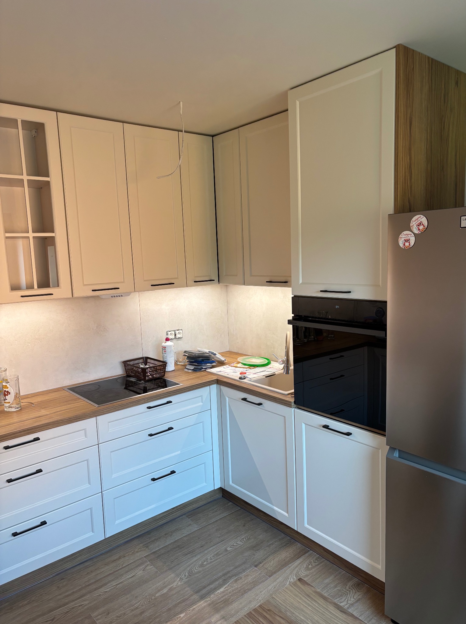 Jasna kuchnia z drewnianym blatem i białymi frontami. Widoczne szafki wiszące i stojące, płyta indukcyjna, piekarnik, lodówka. Uwagę zwraca drewniane wykończenie boku szafki.