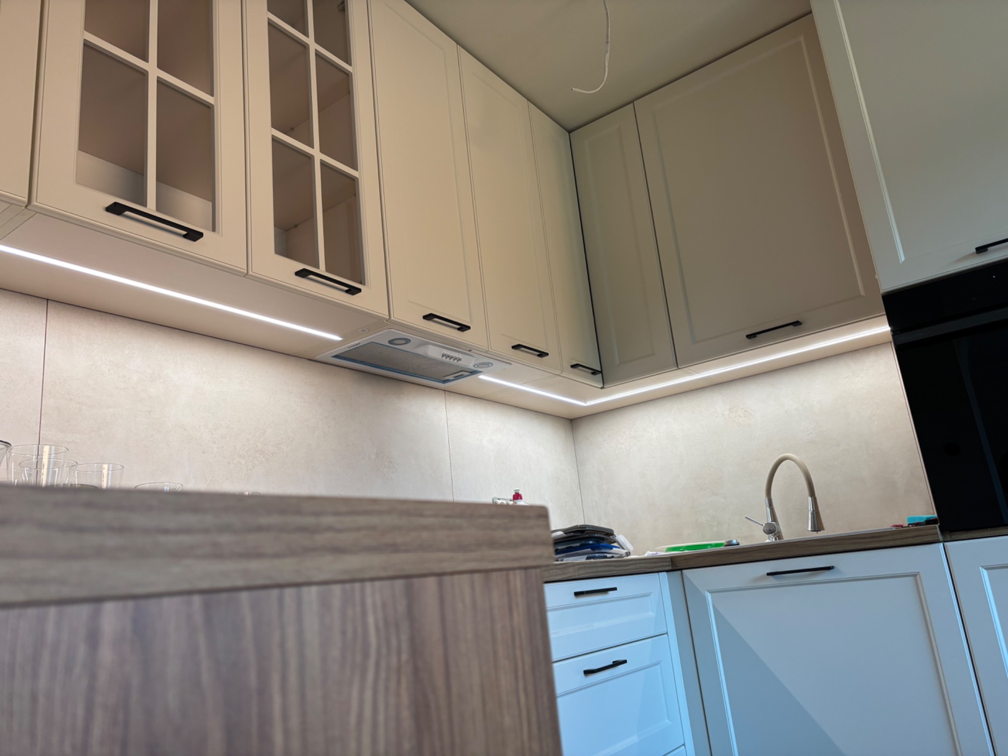 Jasna kuchnia z oświetleniem LED pod szafkami. Górne szafki z przeszkleniami, dolne białe z ciemnymi uchwytami. Ujęcie z dołu, widoczny fragment blatu.