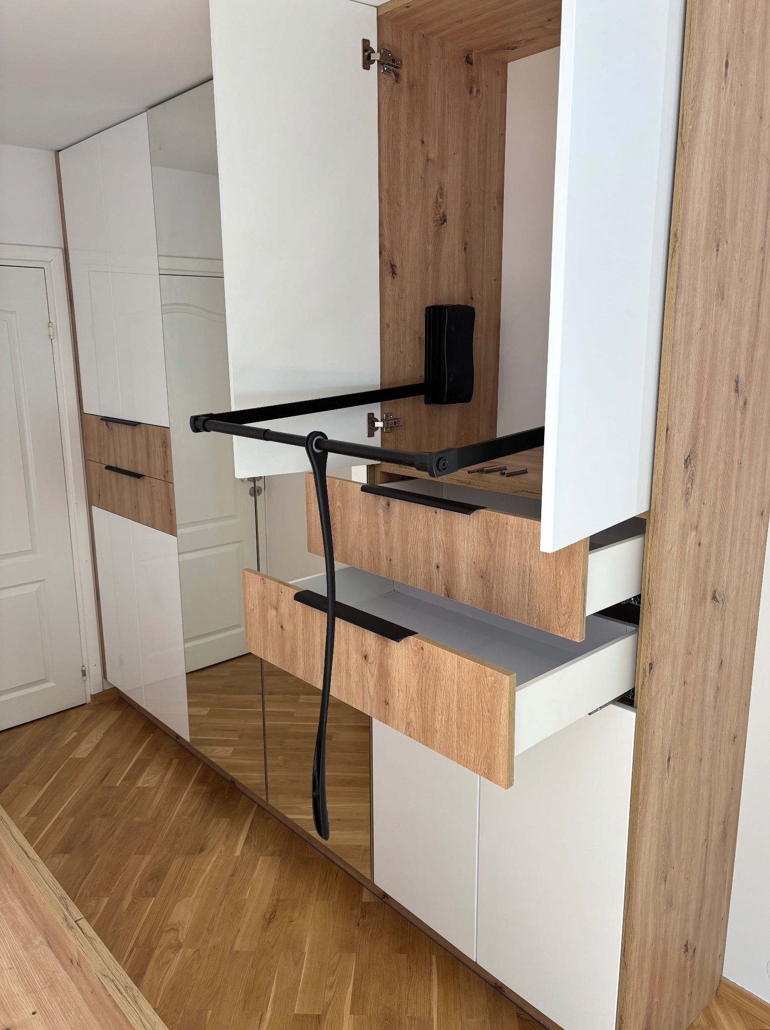 Biało-drewniana szafa wnękowa z wysuwanymi szufladami i drążkiem na ubrania. Widoczne detale wykończenia i mechanizmy. Parkiet na podłodze.