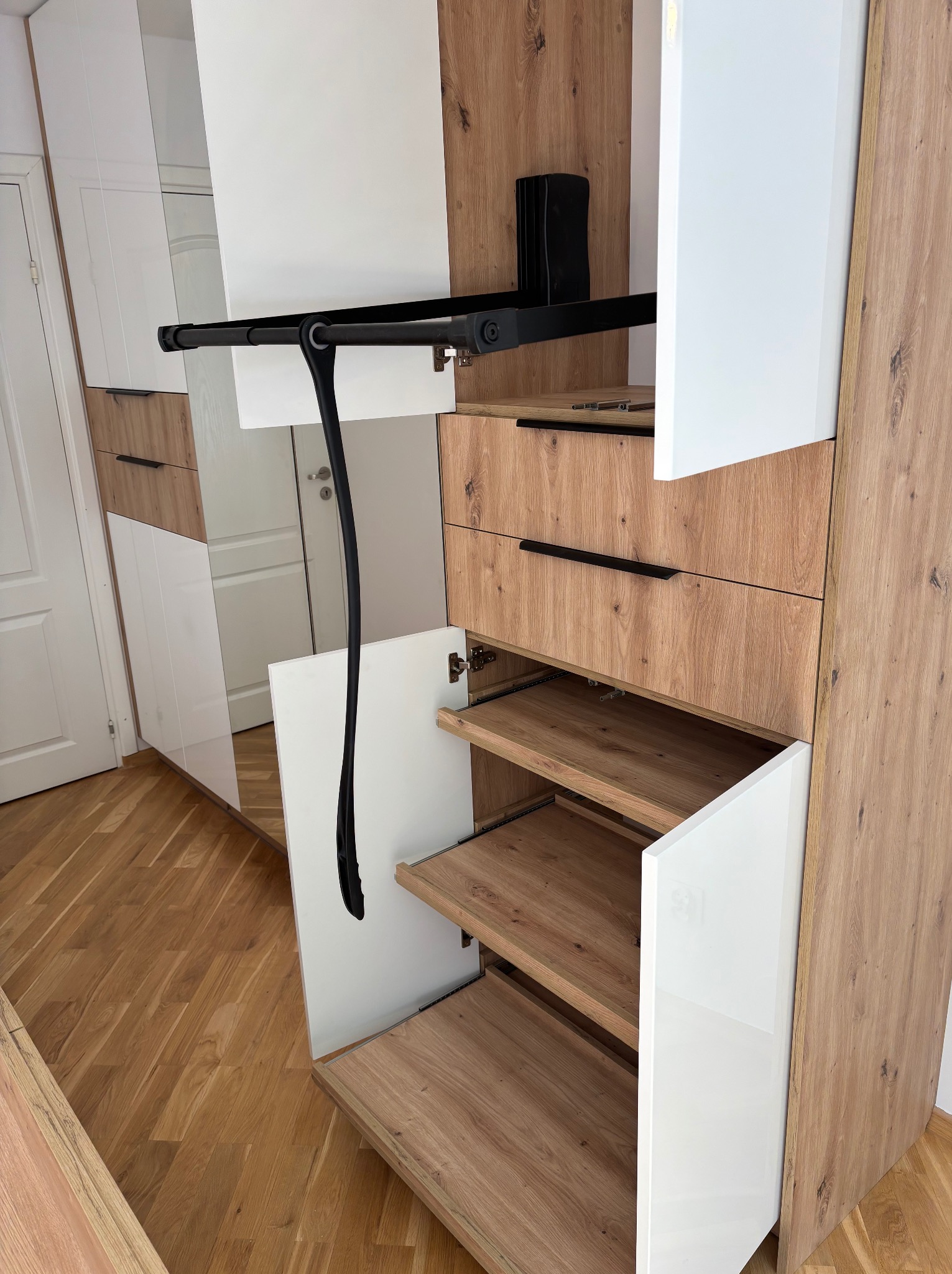 Szafa na wymiar z otwartymi półkami i wysuwanym drążkiem na ubrania. Fronty w kolorze białym i drewnopodobnym. Ujęcie z bliska, widoczny detal.