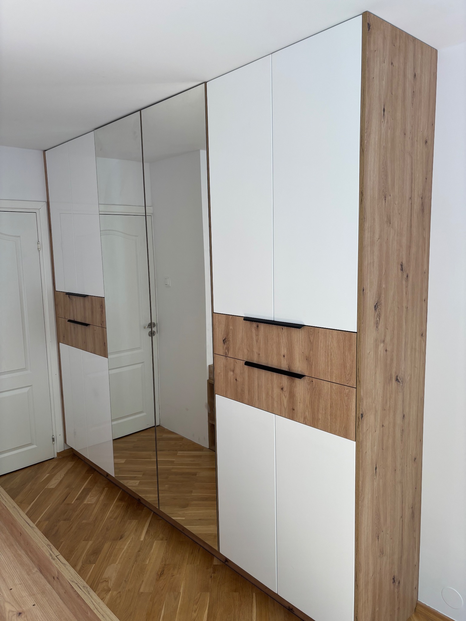 Nowoczesna szafa wnękowa z lustrzanymi drzwiami, biało-drewniane fronty, czarne uchwyty, parkiet na podłodze. Minimalistyczny design.