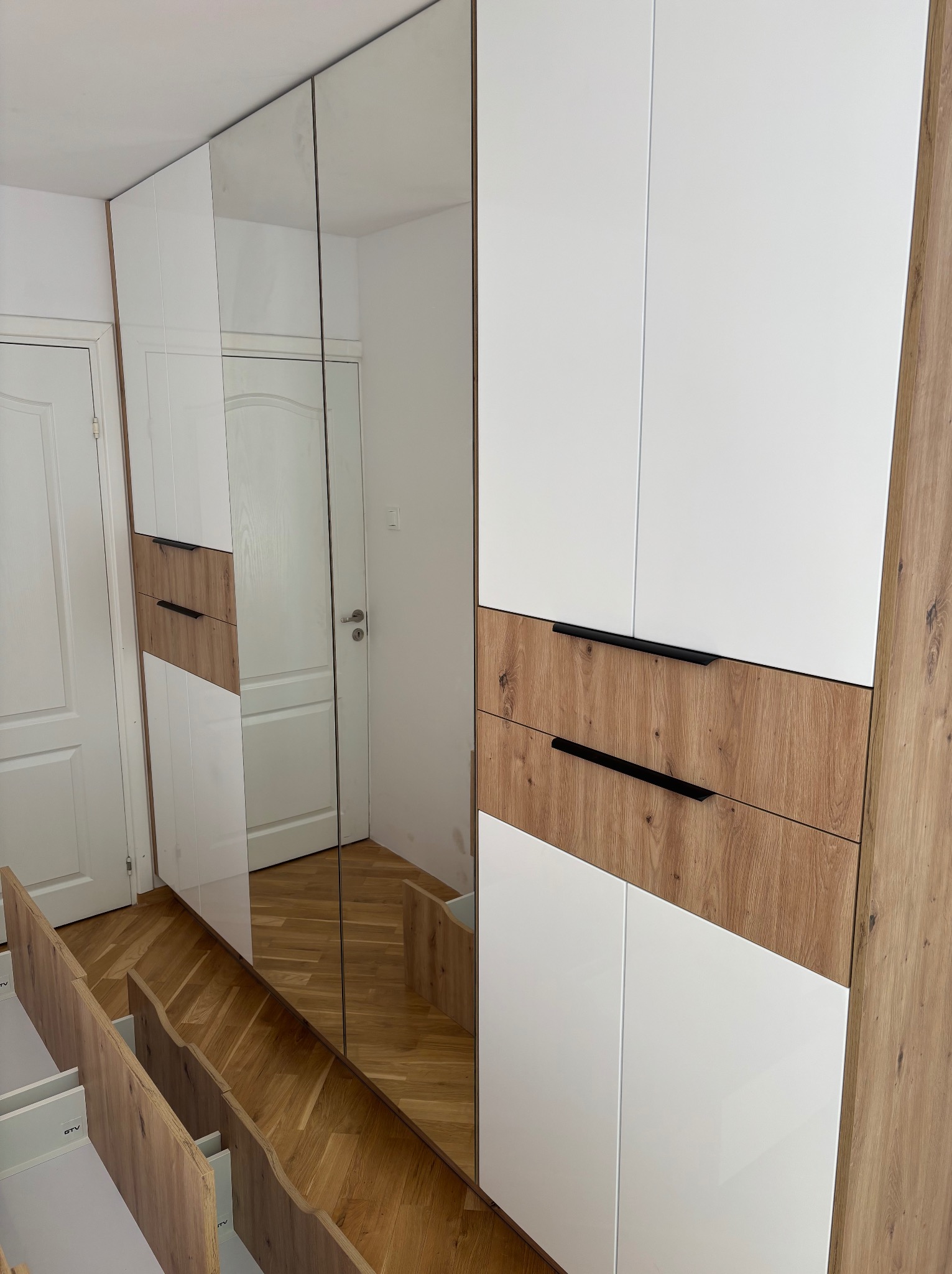 Nowoczesna szafa wnękowa z lustrami i drewnianymi akcentami. Otwarta szuflada ukazuje wnętrze. Jasne wnętrze pomieszczenia odbija się w lustrzanych drzwiach.