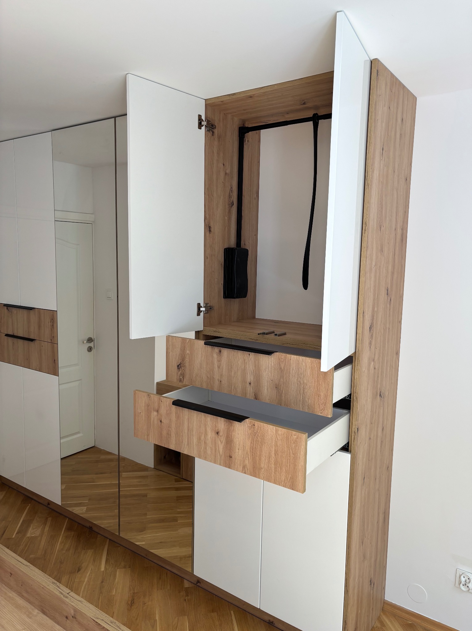 Nowoczesna szafa wnękowa z lustrzanymi drzwiami, otwartymi szufladami i drzwiczkami, wykończona w bieli i drewnie, z widocznym wnętrzem szafy i elementami garderoby.