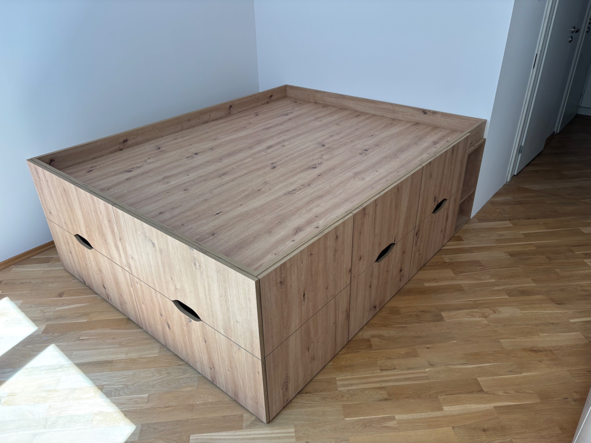 Drewniana zabudowa meblowa z szufladami i otwartą półką, wkomponowana w przestrzeń pokoju, z widocznym parkietem i fragmentem białej ściany.