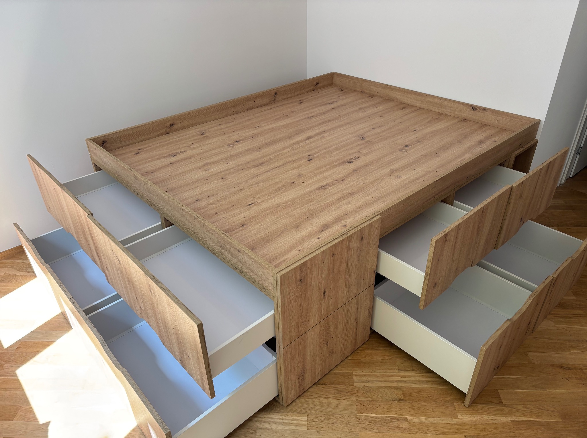 Drewniana platforma z wysuwanymi szufladami, widoczne otwarte białe wnętrza szuflad, na podłodze parkiet. Nietypowa konstrukcja mebla.