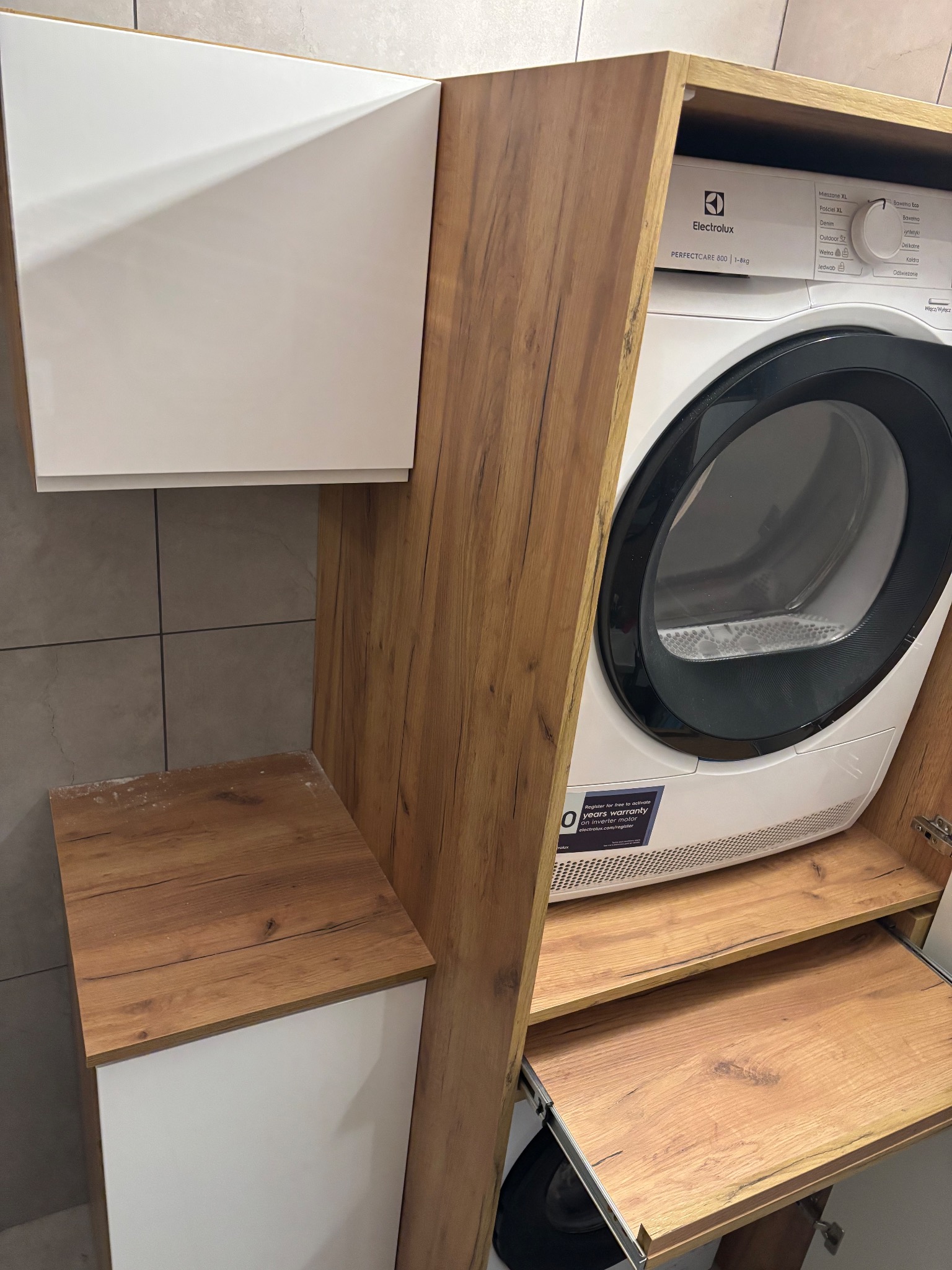 Biała suszarka Electrolux w zabudowie z drewnopodobnej płyty meblowej z wysuwaną półką. Ujęcie z bliska, widoczne detale wykończenia i faktura materiałów.