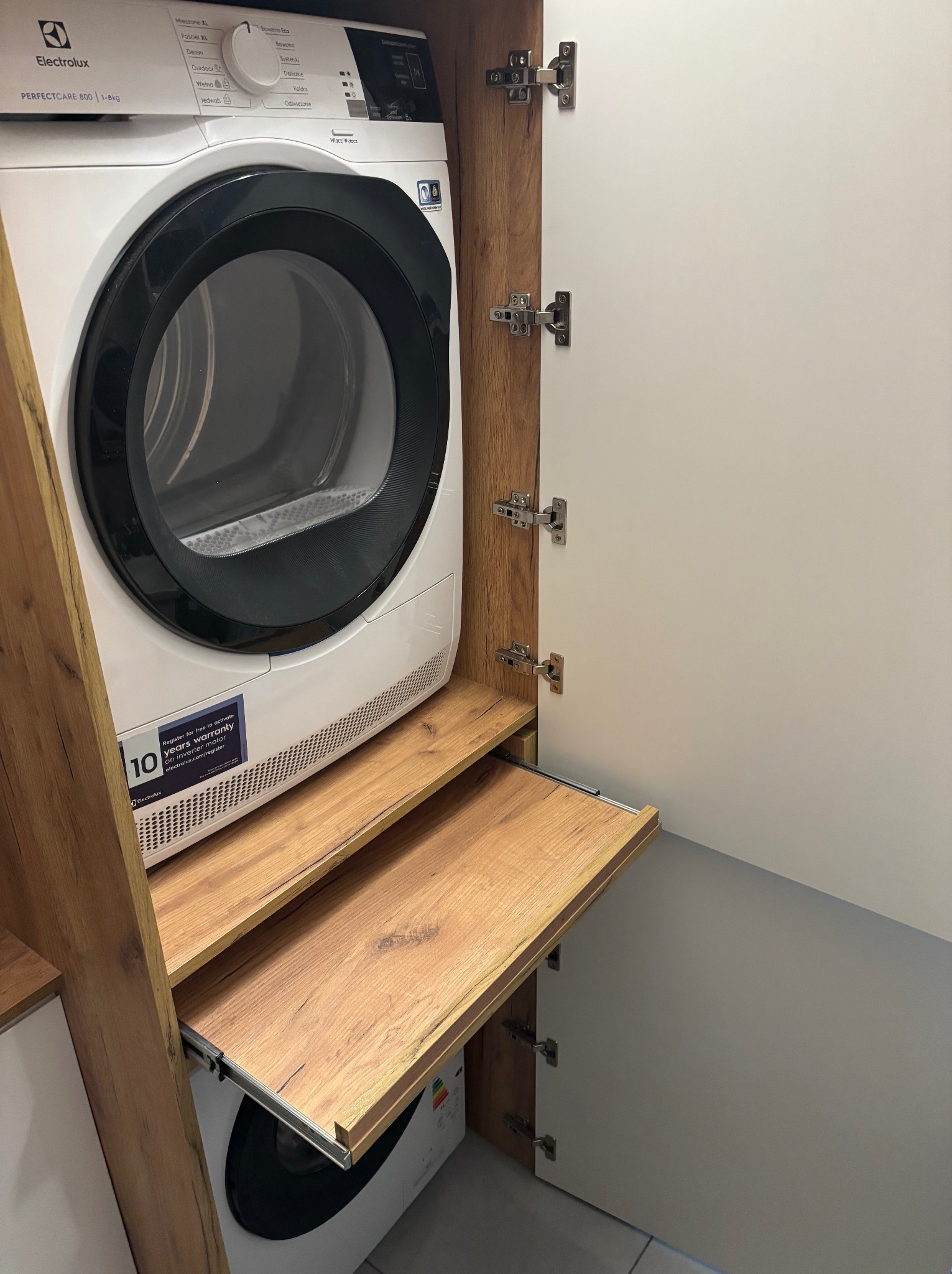 Pralka i suszarka w zabudowie meblowej z drewnianymi elementami i wysuwanym blatem. Białe fronty szafek z widocznymi zawiasami. Nowoczesne rozwiązanie do małej łazienki.