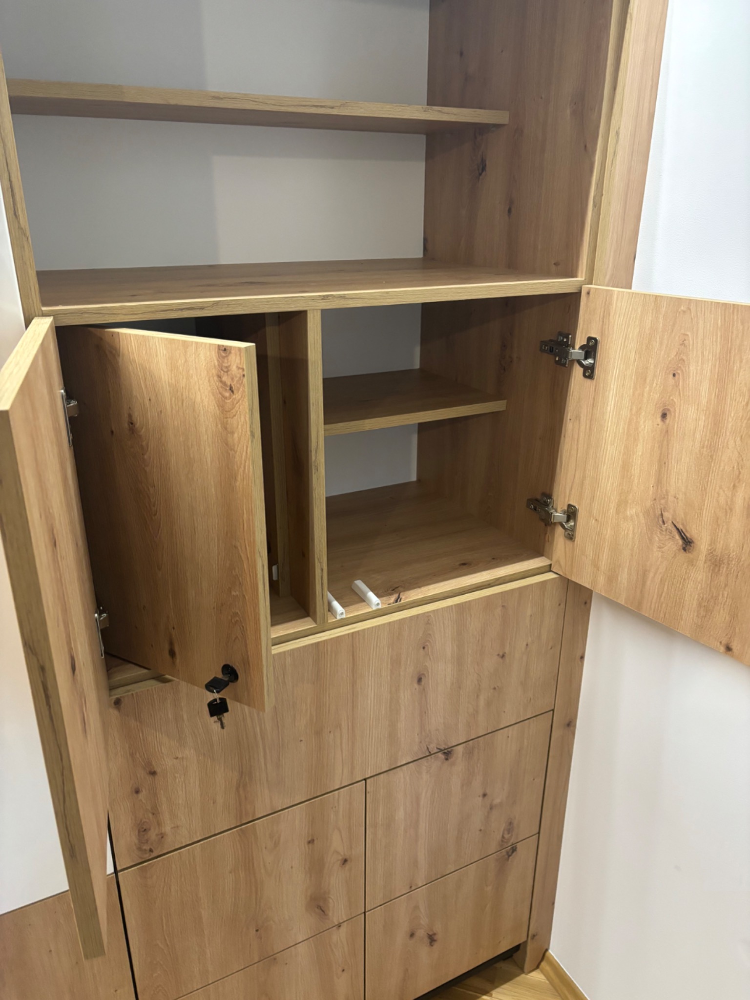 Drewniana szafa z otwartymi drzwiami, widoczne półki i szuflady. Naturalny odcień drewna z wyraźnym usłojeniem. Kluczyk w zamku. Minimalistyczny design.