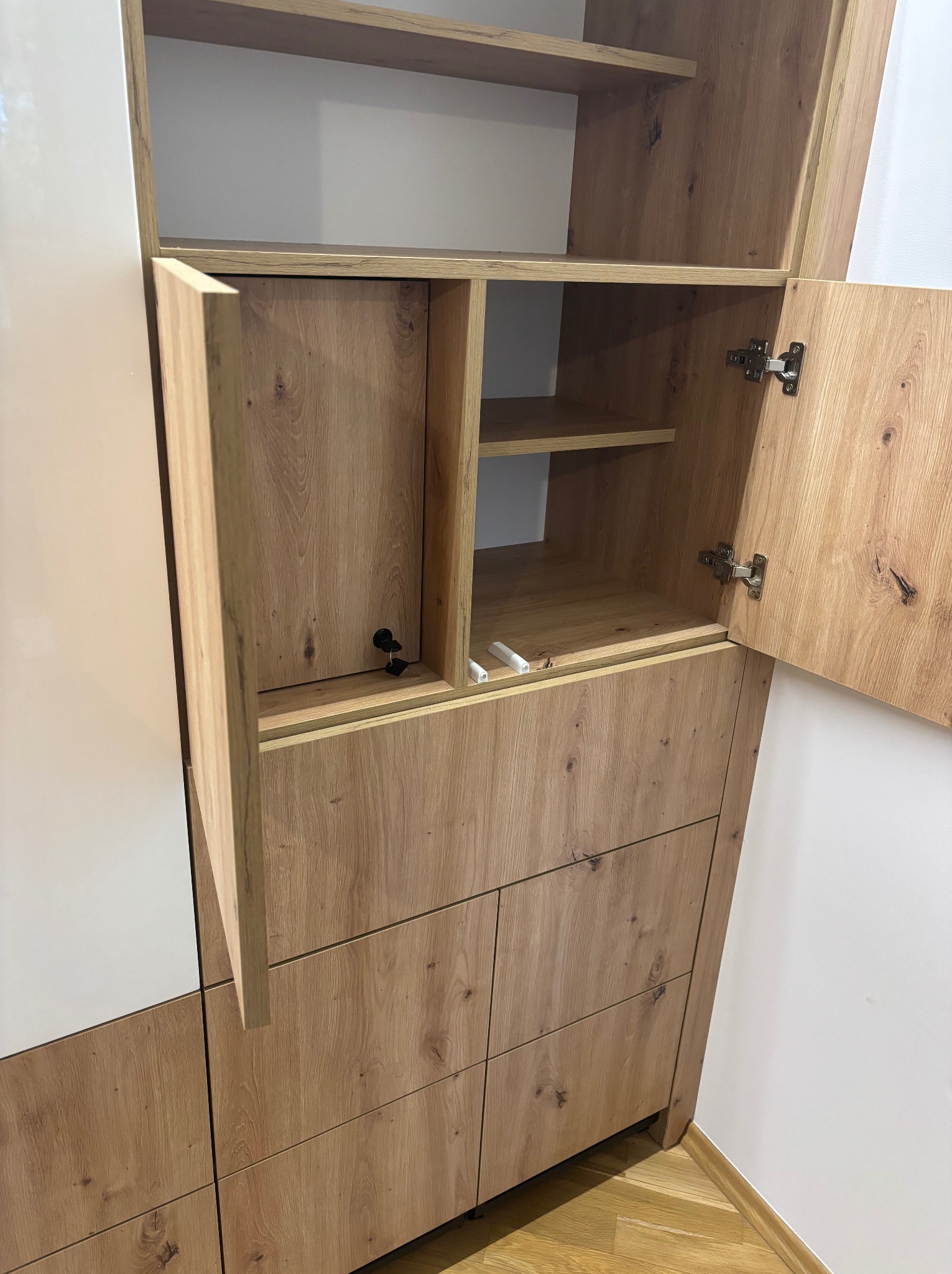 Drewniana szafa z otwartymi drzwiczkami, widoczne półki i szuflady. Fronty w kolorze dębu z białymi elementami. Uwagę przykuwają detale wykończenia i okucia.