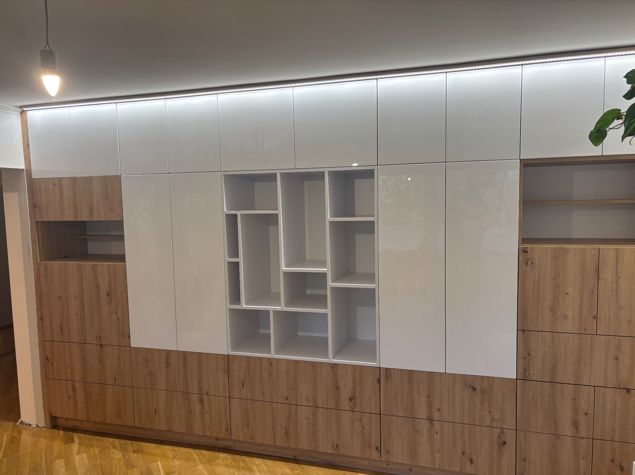 Nowoczesna szafa wnękowa z białymi, lakierowanymi frontami i elementami z drewna. Asymetryczne półki w centralnej części. Oświetlenie LED górne. Parkiet na podłodze.