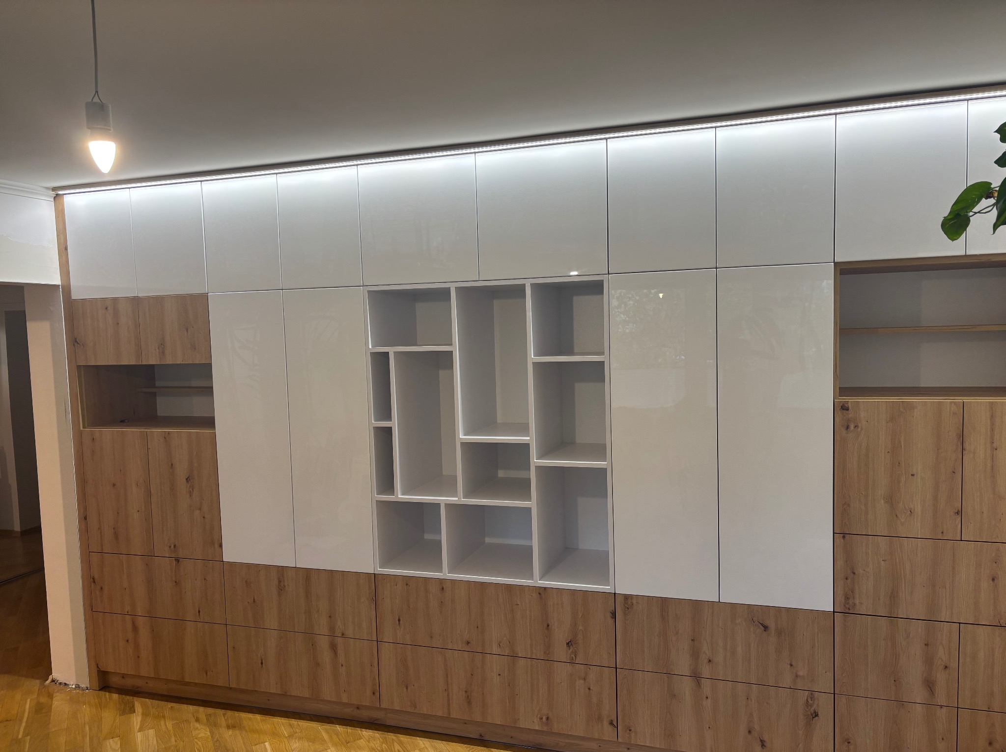 Nowoczesna zabudowa meblowa z białymi, lakierowanymi frontami i drewnopodobnymi panelami. Oświetlenie LED podkreśla geometryczny układ półek i szafek.