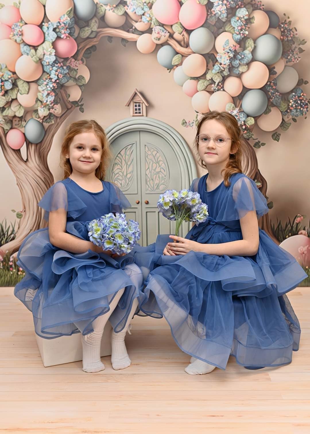 Dwie dziewczynki w błękitnych, tiulowych sukienkach siedzą na tle dekoracji imitującej bajkowy ogród z drzwiami i balonami, trzymając bukieciki niebieskich kwiatów.