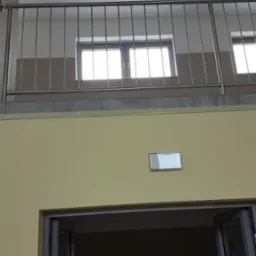 Wewnętrzna balustrada ze stali nierdzewnej z pionowymi prętami zabezpieczająca antresolę, widok z dołu na żółtą ścianę z oświetleniem i otwartym wejściem do pomieszczenia.