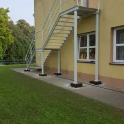 Zewnętrzne metalowe schody ewakuacyjne z balustradą, przymocowane do żółtej ściany budynku, widoczne okna z dekoracjami.