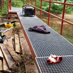 E&P PRO-METAL spółka jawna E.Luty P.Luty - Metalowa kładka nad strumieniem w lesie w trakcie budowy, widoczny agregat prądotwórczy, robocze rękawice i ubranie, drewniane deski oraz narzędzia w żółtym koszyku.