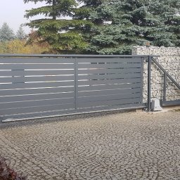 E&P PRO-METAL spółka jawna E.Luty P.Luty - Automatyczna brama przesuwna w kolorze grafitowym z poziomymi panelami, widoczny napęd i ogrodzenie z gabionów wypełnionych kamieniami w tle.