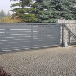 Automatyczna brama przesuwna w kolorze grafitowym z poziomymi panelami, widoczny napęd i ogrodzenie z gabionów wypełnionych kamieniami w tle.