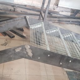 E&P PRO-METAL spółka jawna E.Luty P.Luty - Surowe, metalowe schody z kratowymi stopniami w trakcie montażu, widoczne ślady spawania i niedoskonałości wykończenia, na podłodze leżą metalowe profile.