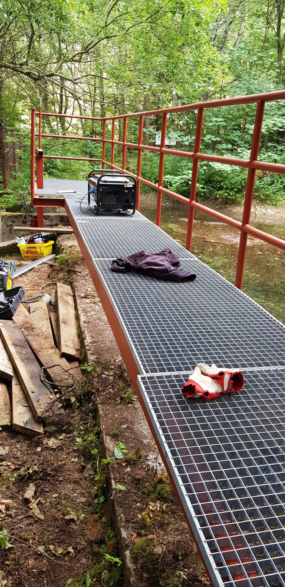 Metalowa kładka nad strumieniem w lesie w trakcie budowy, widoczny agregat prądotwórczy, robocze rękawice i ubranie, drewniane deski oraz narzędzia w żółtym koszyku.
