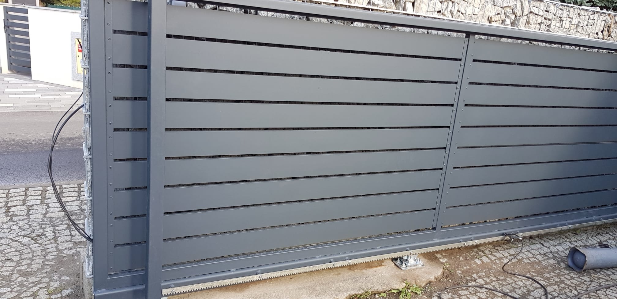 Szara, przesuwna brama wjazdowa z poziomymi panelami, widoczne elementy automatyki i napędu, tło z kamiennego muru.