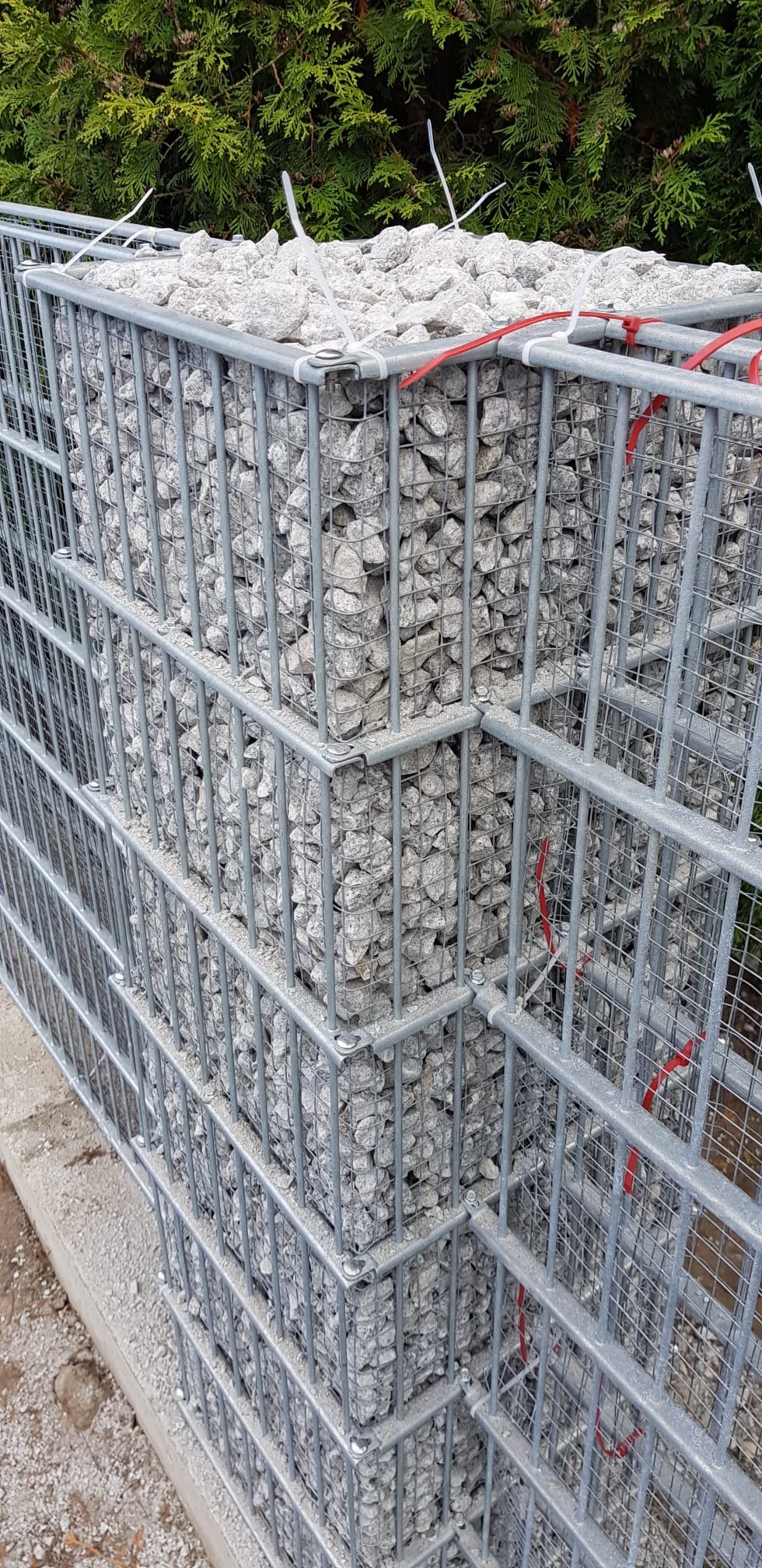 Gabion z szarego kamienia, zabezpieczony metalową siatką, z widocznymi białymi i czerwonymi opaskami zaciskowymi, na tle zielonych krzewów.
