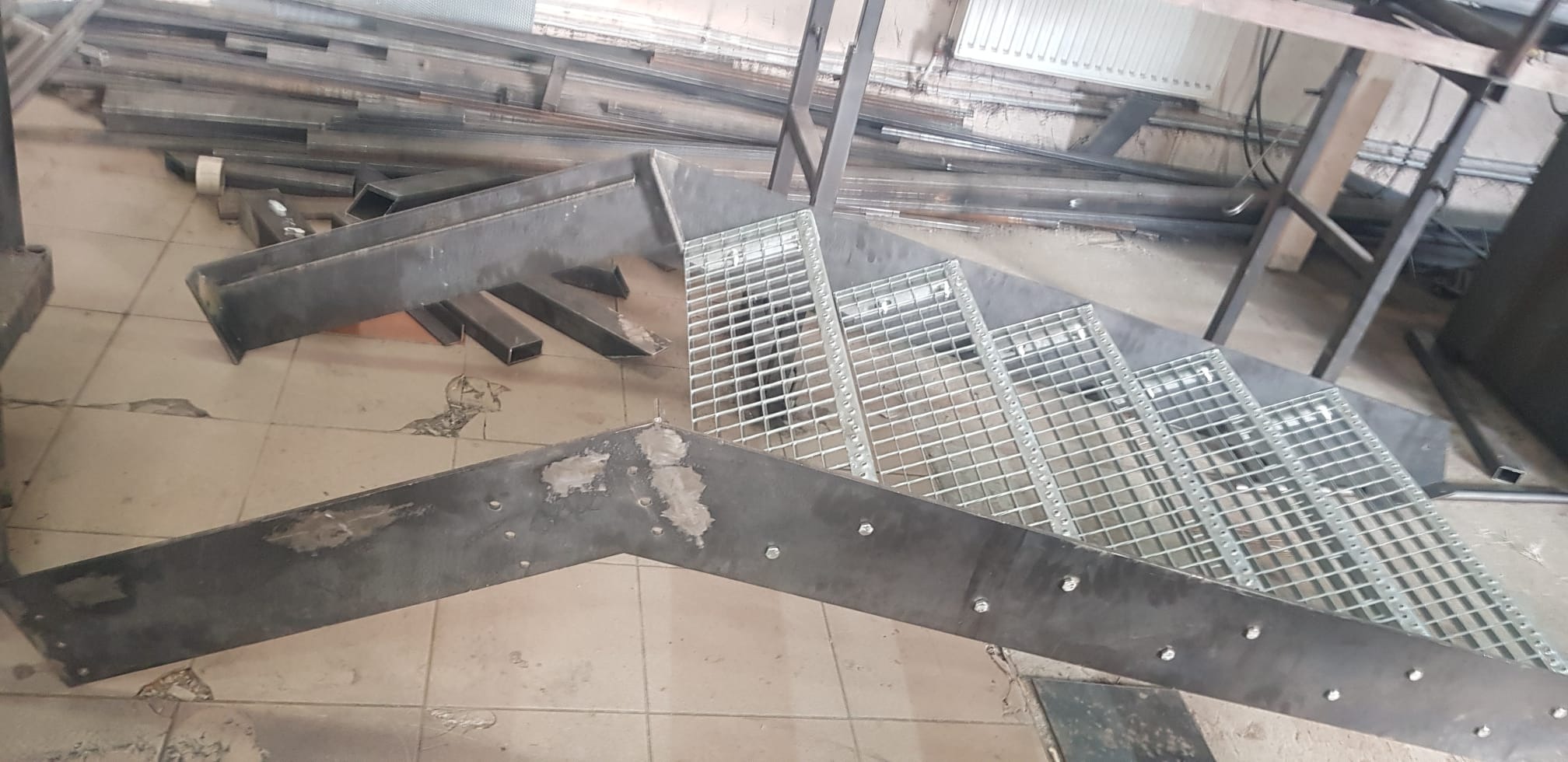 Surowe, metalowe schody z kratowymi stopniami w trakcie montażu, widoczne ślady spawania i niedoskonałości wykończenia, na podłodze leżą metalowe profile.