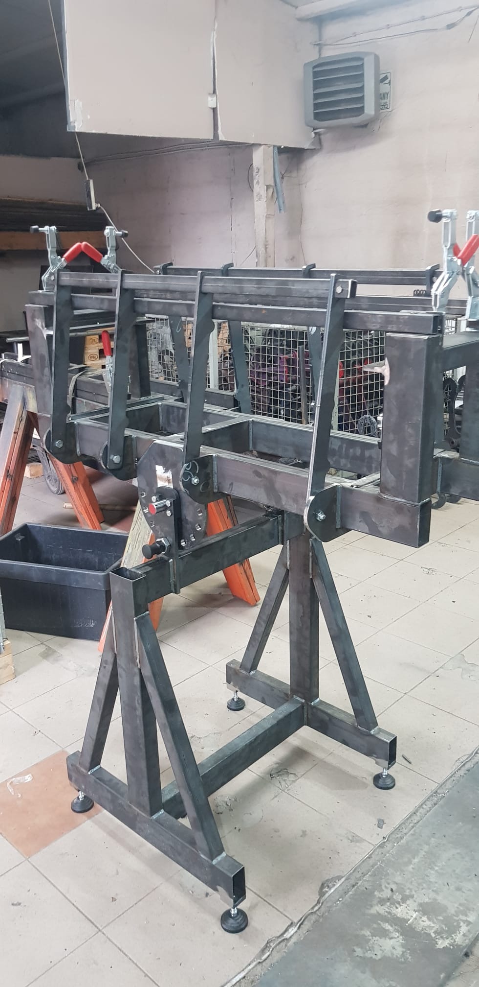 Surowa, metalowa konstrukcja spawana na regulowanych nóżkach, z klamrami zaciskowymi, prawdopodobnie w warsztacie, na tle ściany z grzejnikiem.