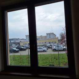 FPMULTISERWIS Fortunata Piec - Widok z okna na parking z samochodami, zieleń i budynki w pochmurny dzień, rama okienna w ciemnym kolorze, grzejnik pod parapetem.