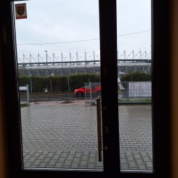 FPMULTISERWIS Fortunata Piec - Widok przez otwarte drzwi na mokrą kostkę brukową, ogrodzenie, żywopłot, czerwony samochód i stadion w tle, pochmurne niebo.