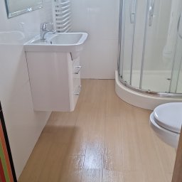 FPMULTISERWIS Fortunata Piec - Jasna łazienka z kabiną prysznicową, umywalką z szafką i grzejnikiem drabinkowym pod oknem, wykończona płytkami imitującymi drewno.