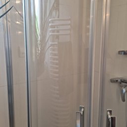 FPMULTISERWIS Fortunata Piec - Szklana kabina prysznicowa z chromowanymi uchwytami, widoczny grzejnik drabinkowy i roślina w tle, odbicie światła na szkle.