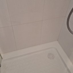 FPMULTISERWIS Fortunata Piec - Biały brodzik kabiny prysznicowej z widocznymi resztkami piany, fragment szklanej ściany kabiny i chromowany drążek prysznicowy na tle białych, kwadratowych płytek ściennych.