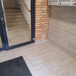 FPMULTISERWIS Fortunata Piec - Wnętrze klatki schodowej z widocznymi schodami za przeszklonymi drzwiami, ceglana kolumna i rzędy skrzynek pocztowych nad marmurową posadzką.