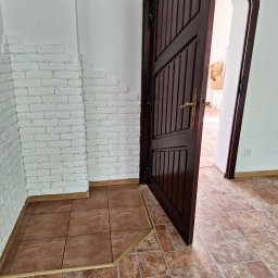 FPMULTISERWIS Fortunata Piec - Korytarz z brązową podłogą z płytek, białą ścianą z dekoracyjną cegłą i otwartymi, ciemnymi drzwiami z mosiężną klamką.