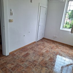 FPMULTISERWIS Fortunata Piec - Puste wnętrze pokoju z brązową terakotą, białymi ścianami, oknem z kratami i klimatyzatorem na ścianie.