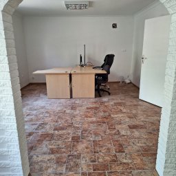 FPMULTISERWIS Fortunata Piec - Biuro z brązową podłogą z płytek, biurkiem, krzesłem i komputerem, widoczne przez łuk z białej cegły.