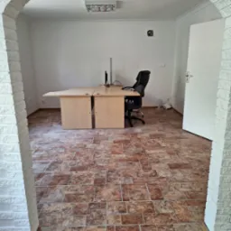 Biuro z brązową podłogą z płytek, biurkiem, krzesłem i komputerem, widoczne przez łuk z białej cegły.